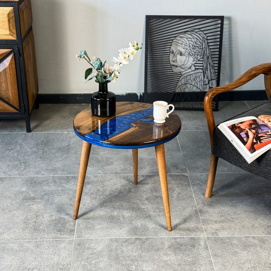 Round walnut side table with a blue epoxy river and three tapered legs beside an armchair