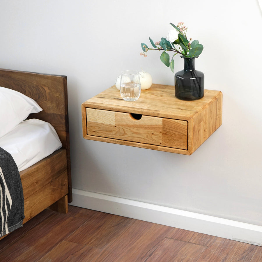 Solid oak floating nightstand with a drawer mounted on the wall beside a bed, with a glass and vase on top