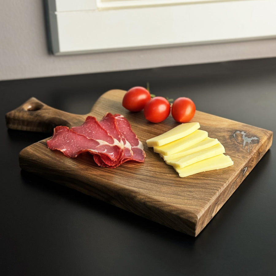 Walnut chopping board with a paddle handle at the back left, angled view with sliced cheese on a black counter