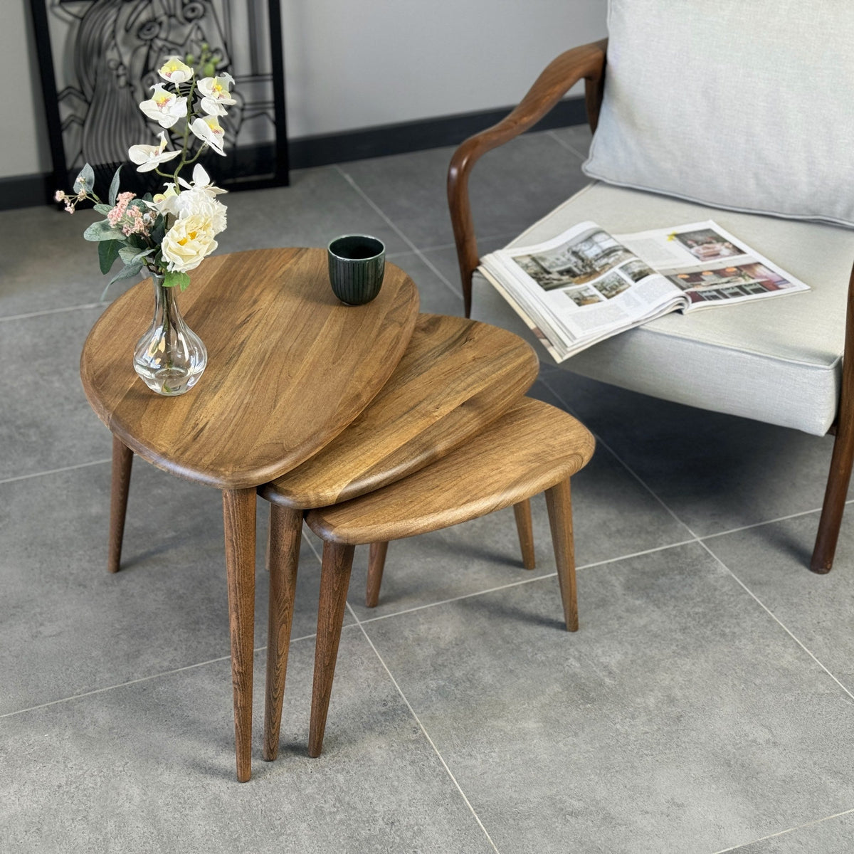 Three walnut nesting tables with pebble-shaped tops and tapered legs, fanned out on a grey tile floor beside an armchair