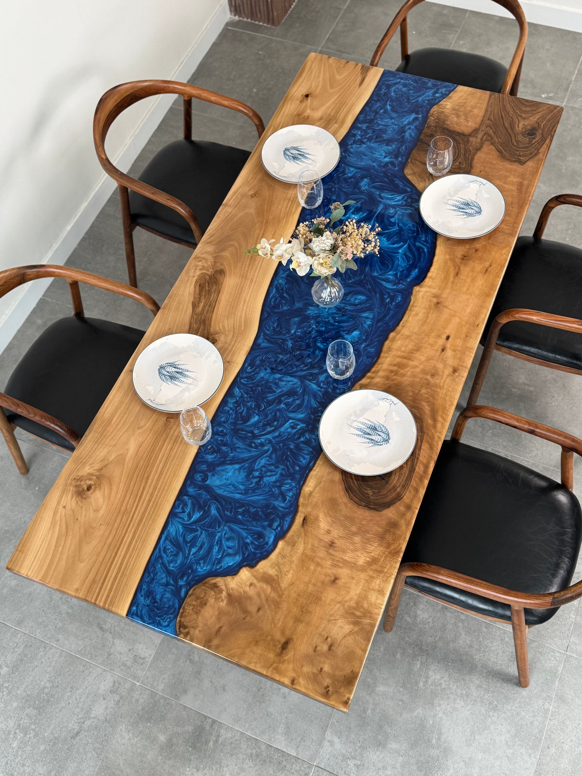 Top-down view of a walnut dining table with a wide, swirling blue epoxy river down the middle, set with white plates and glasses and surrounded by wooden armchairs