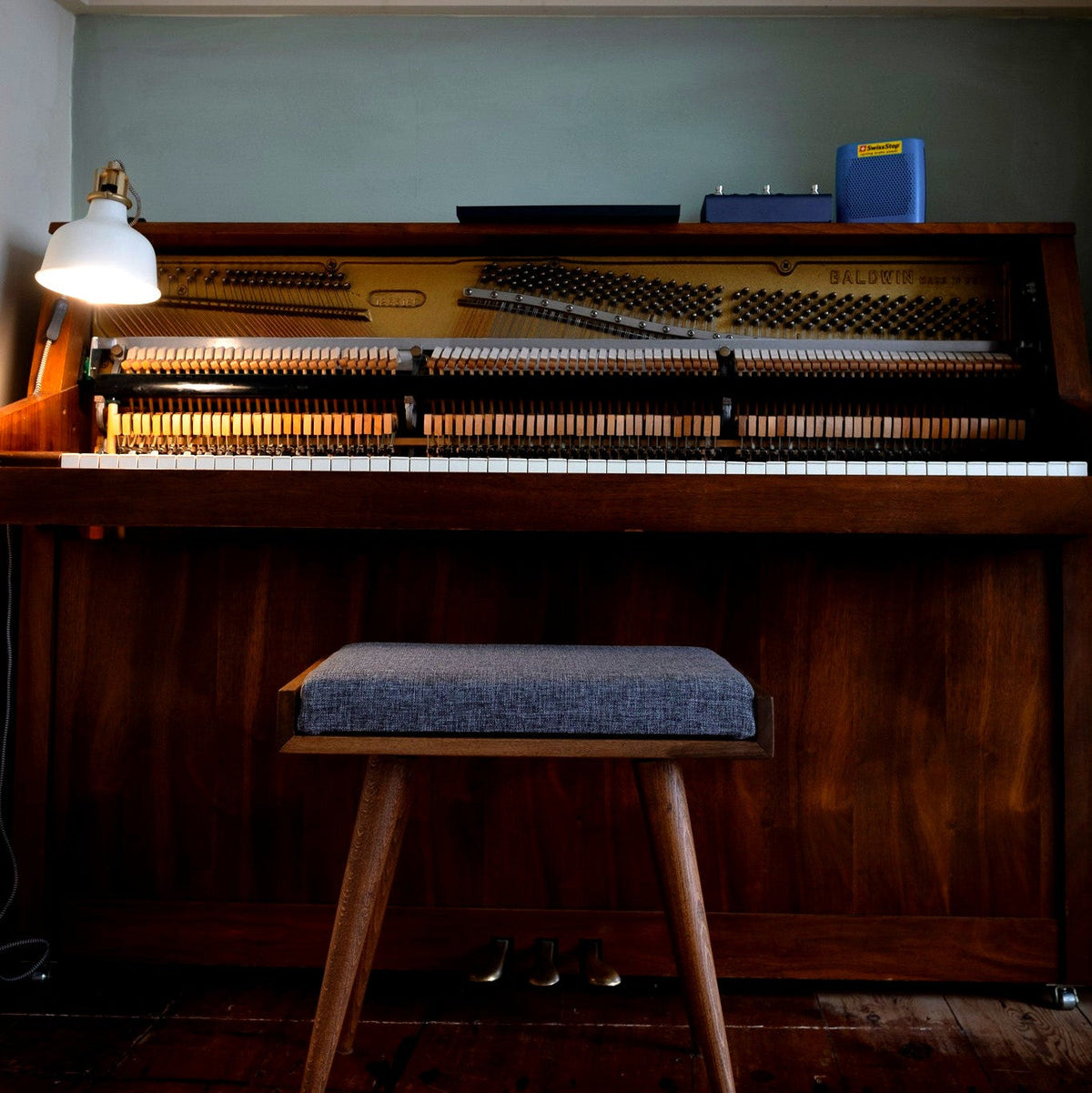 Walnut piano bench with a grey woven cushion in front of an upright piano with its top open, lit by a white clamp lamp