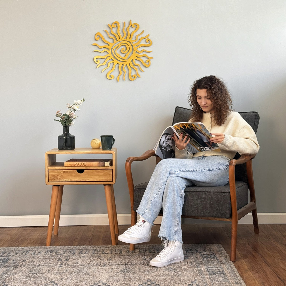 Oak nightstand with drawer and cubby beside an armchair, wide view with a seated reader