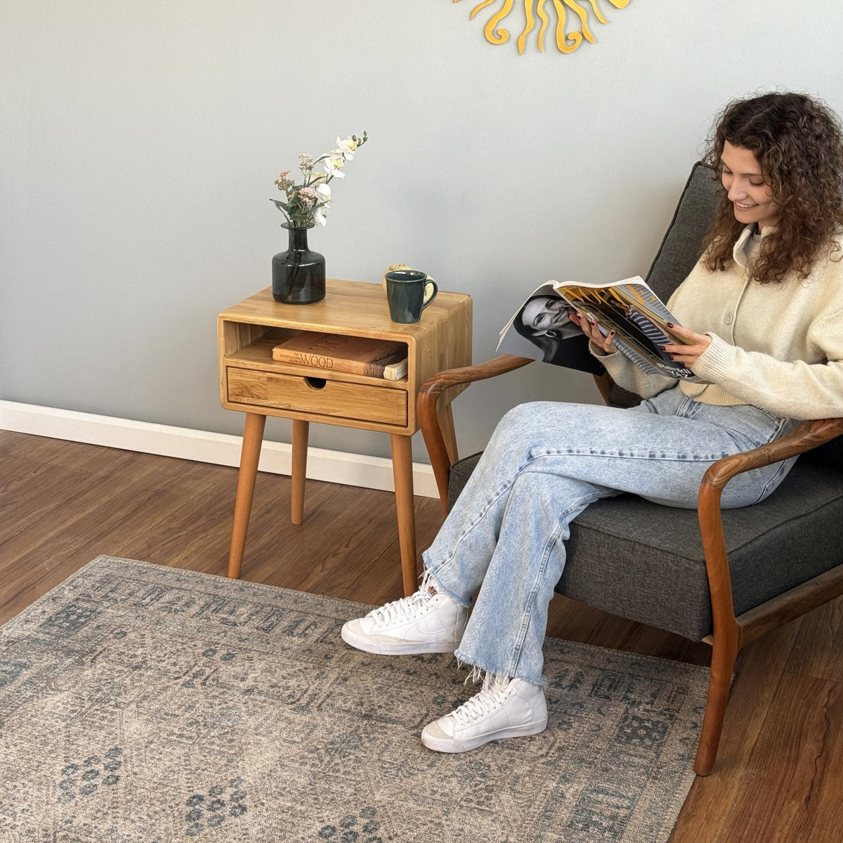 Oak nightstand with open cubby and drawer beside an armchair on a patterned rug, mid-distance view