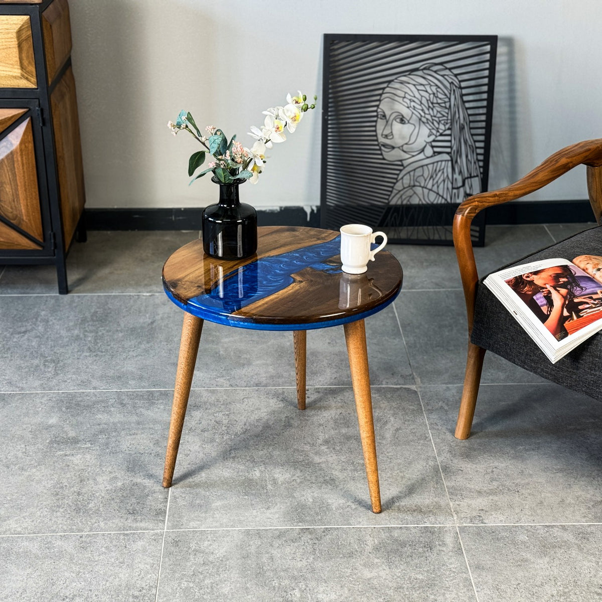 Round walnut side table with a blue epoxy river and three tapered legs beside an armchair