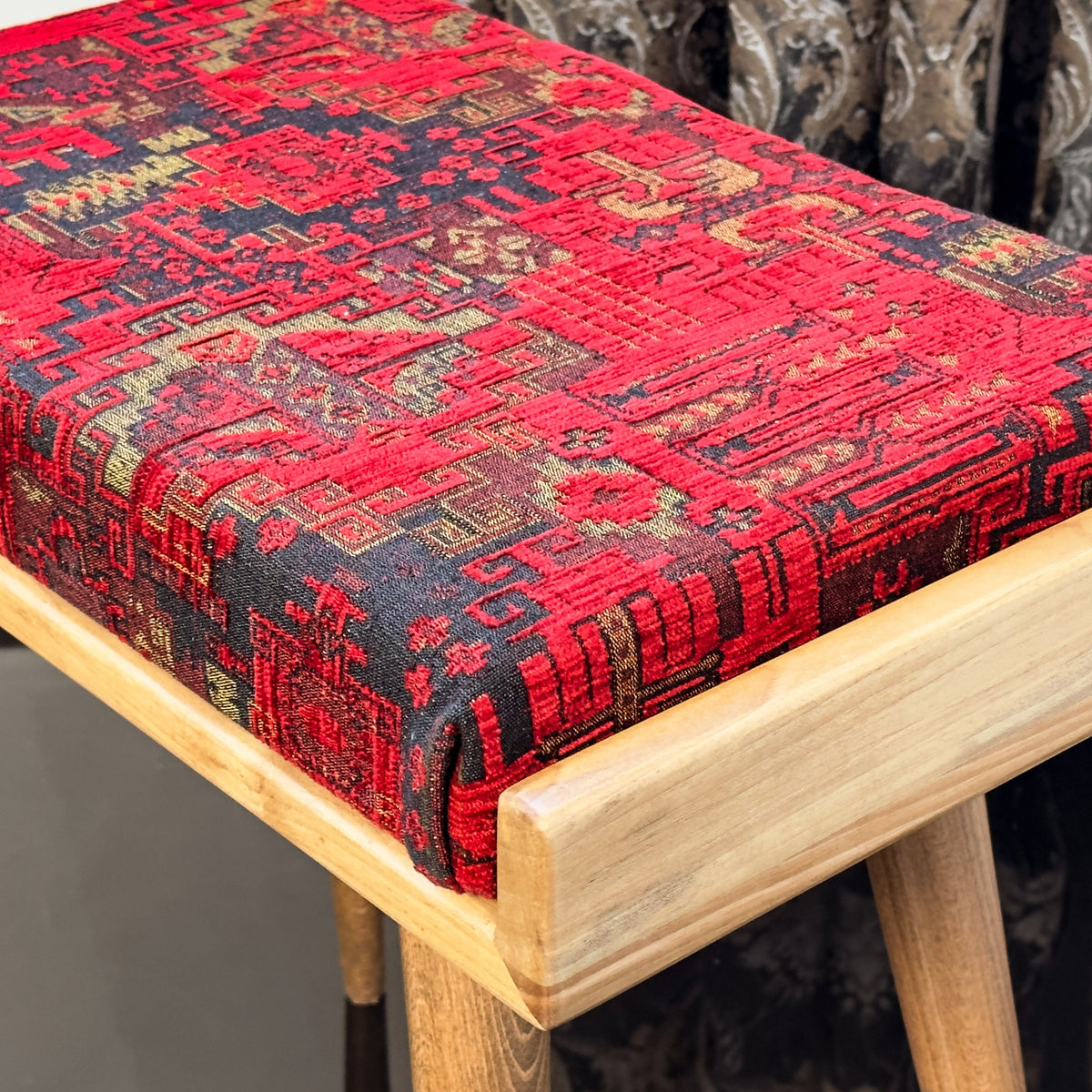 Close-up of the red patterned fabric cushion and the light wooden frame corner of the piano bench