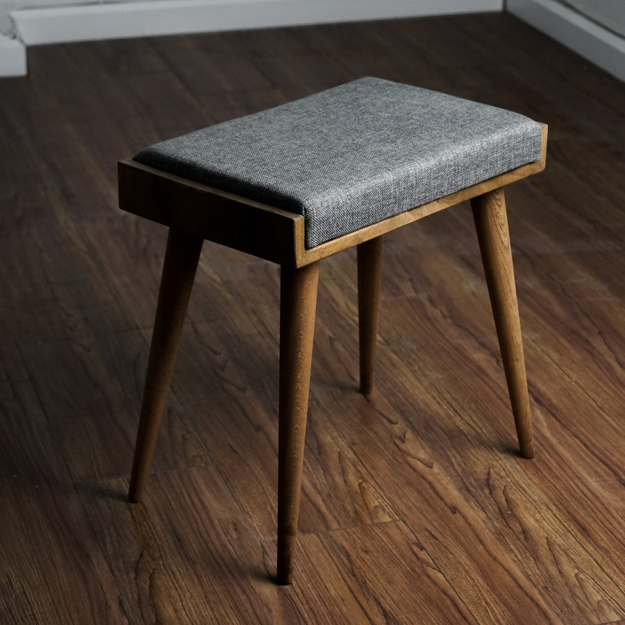 Walnut piano bench with a gray fabric cushion and four tapered legs on a wood floor