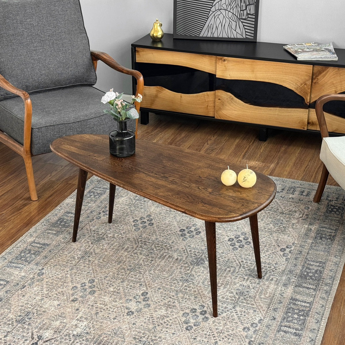 Solid oak oval coffee table with dark tapered legs on a patterned rug, three-quarter view beside a gray armchair