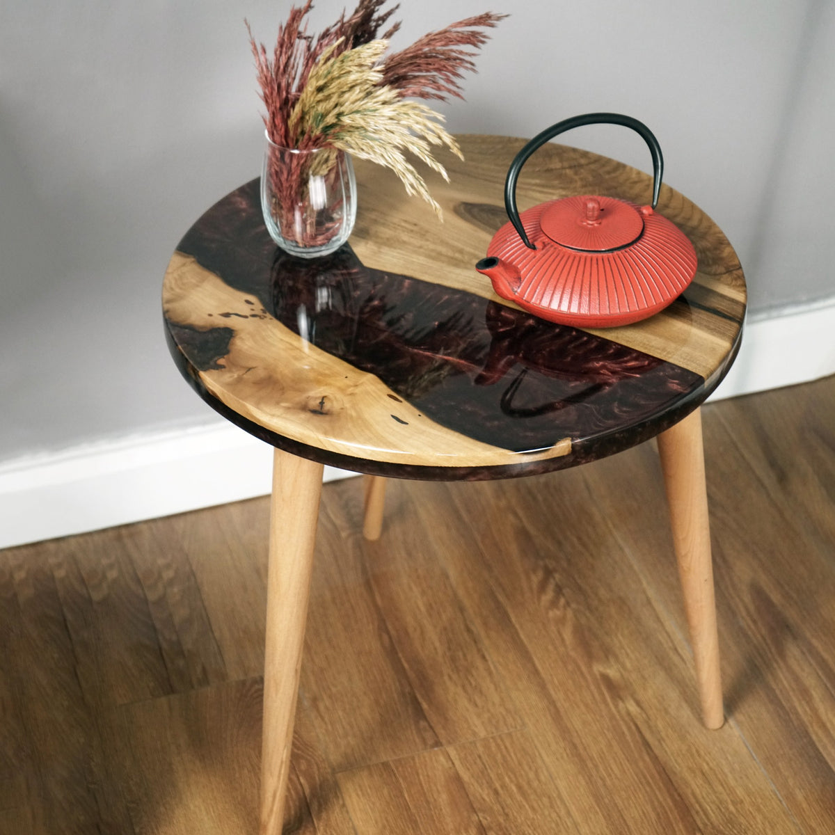 Maroon epoxy river round side table with a live edge walnut top on three tapered legs against a gray wall