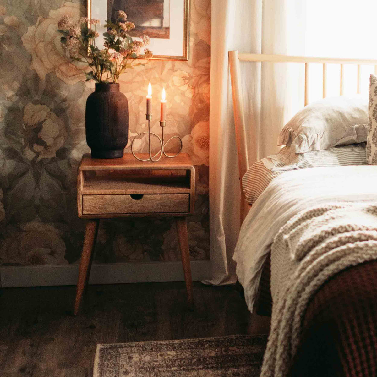Walnut nightstand with shelf and drawer by a bed with a knit throw, candles lit on top