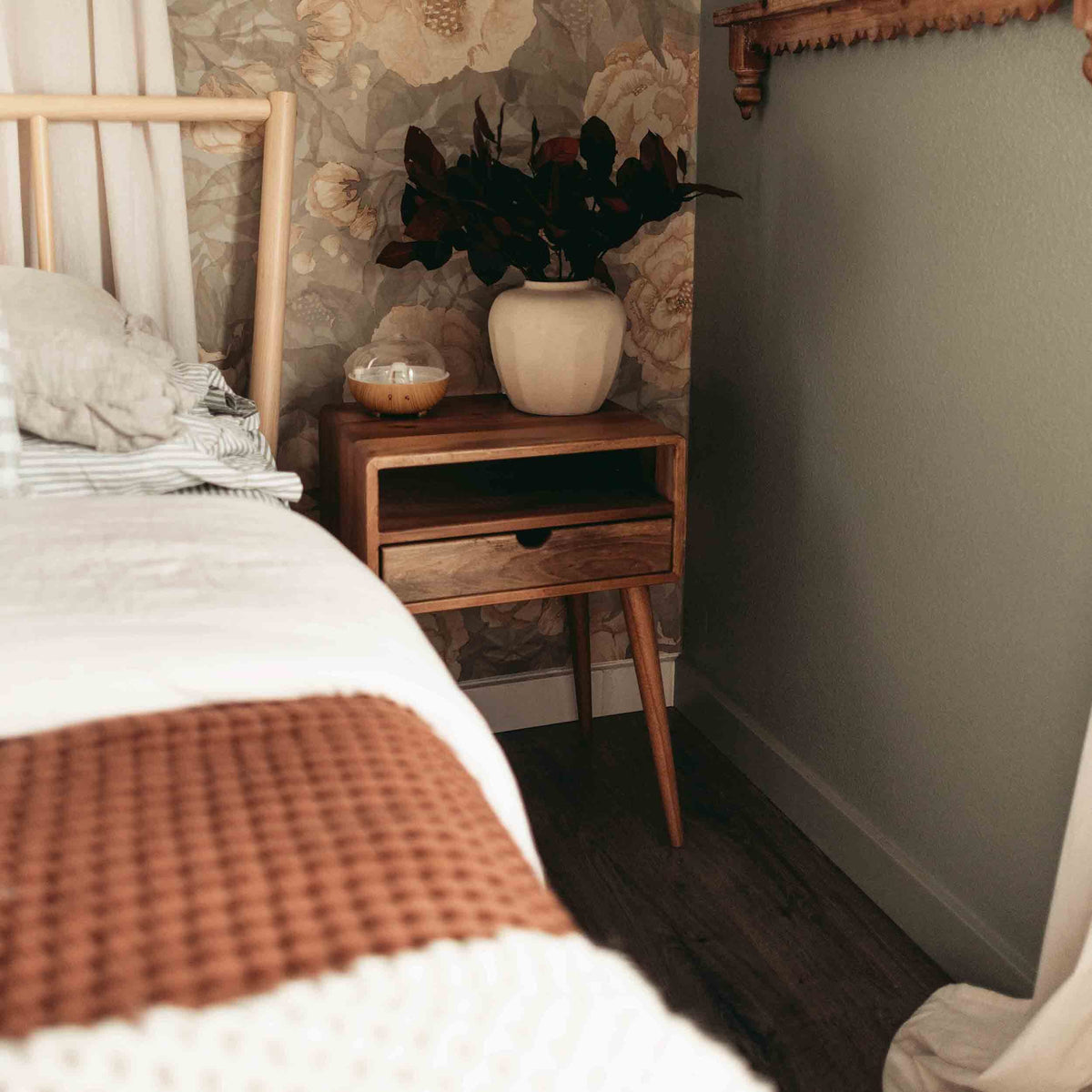 Walnut nightstand with open shelf and drawer by a bed, holding a cream vase with dark leaves
