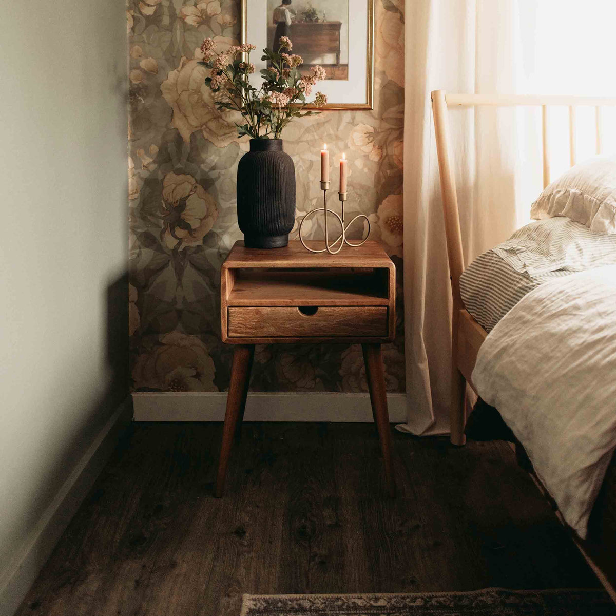 Walnut nightstand with open shelf and drawer next to the bed and a floral wallpaper wall