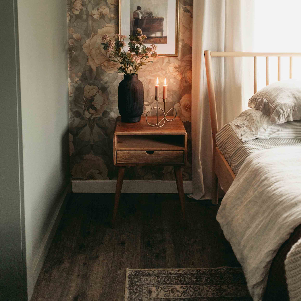 Walnut nightstand with open shelf and drawer in a narrow bedroom corner beside the bed, a lit candle on top