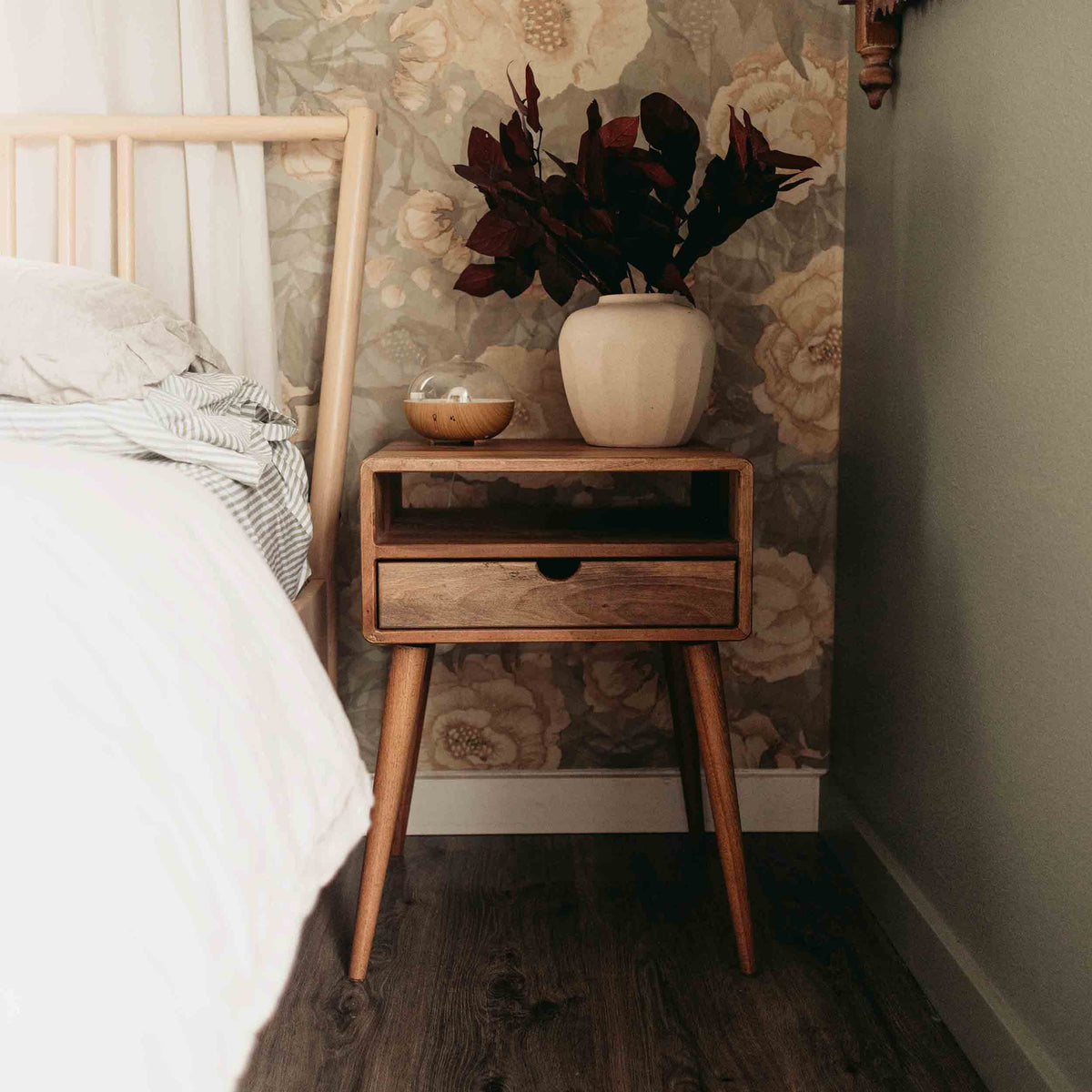 Walnut nightstand with open shelf and drawer on tapered legs, cream vase on top and bed on the left
