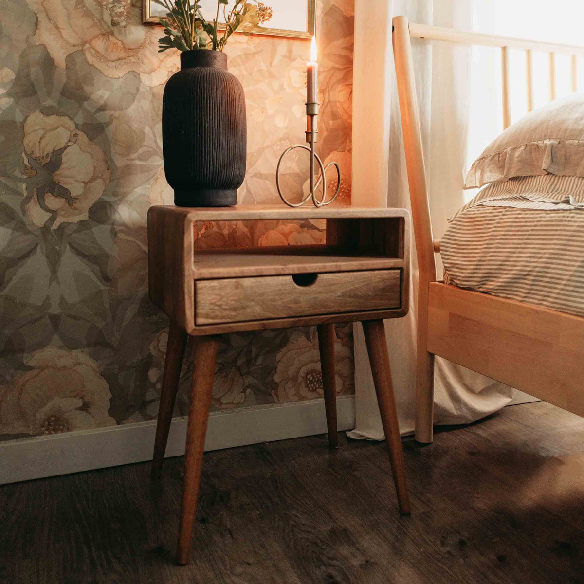 Front view of the walnut nightstand with open shelf, drawer and tapered legs beside a bed