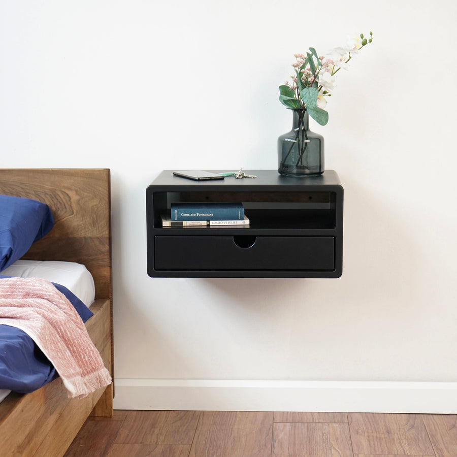 Black wall mounted floating nightstand with a drawer and open shelf beside a wooden bed
