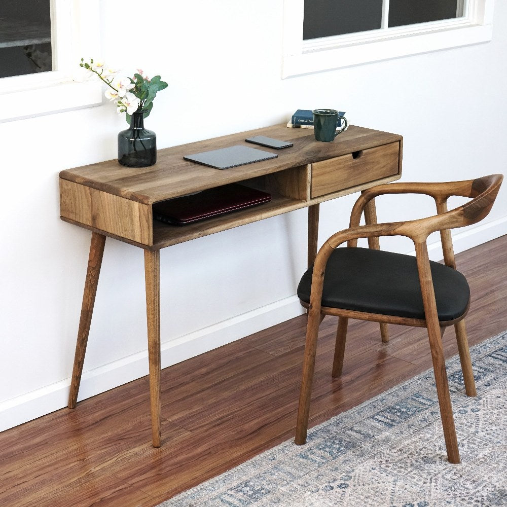 Side angled view of the walnut desk with the drawer closed and a wooden armchair beside it