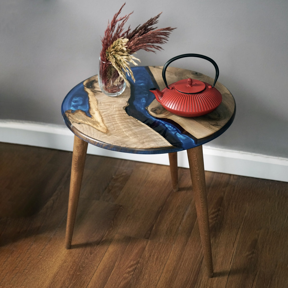 Blue epoxy river side table with a live edge walnut top and a red teapot on a wood floor