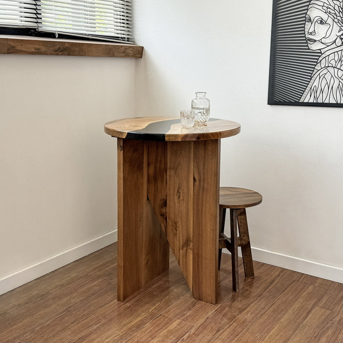 Walnut round bar table with an epoxy river top in a room corner beside a window, with a stool