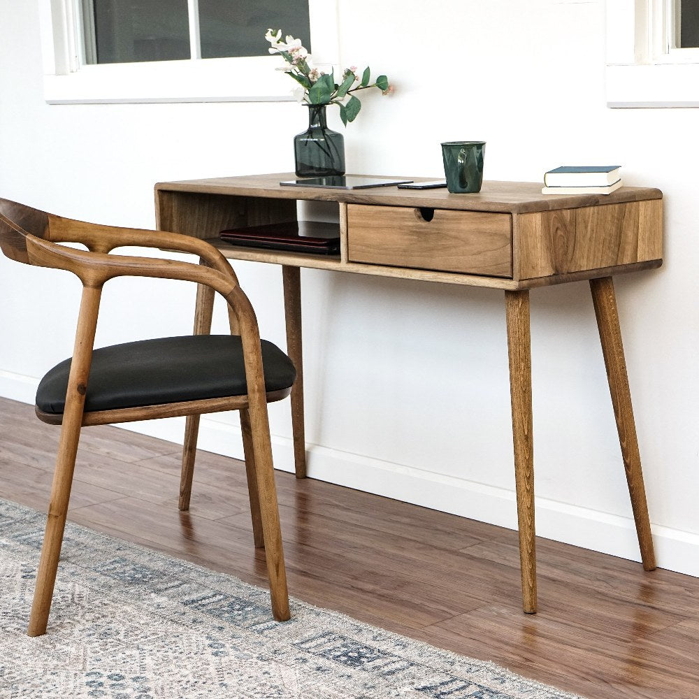 Front view of the walnut desk with the drawer closed, armchair at the left and a window behind