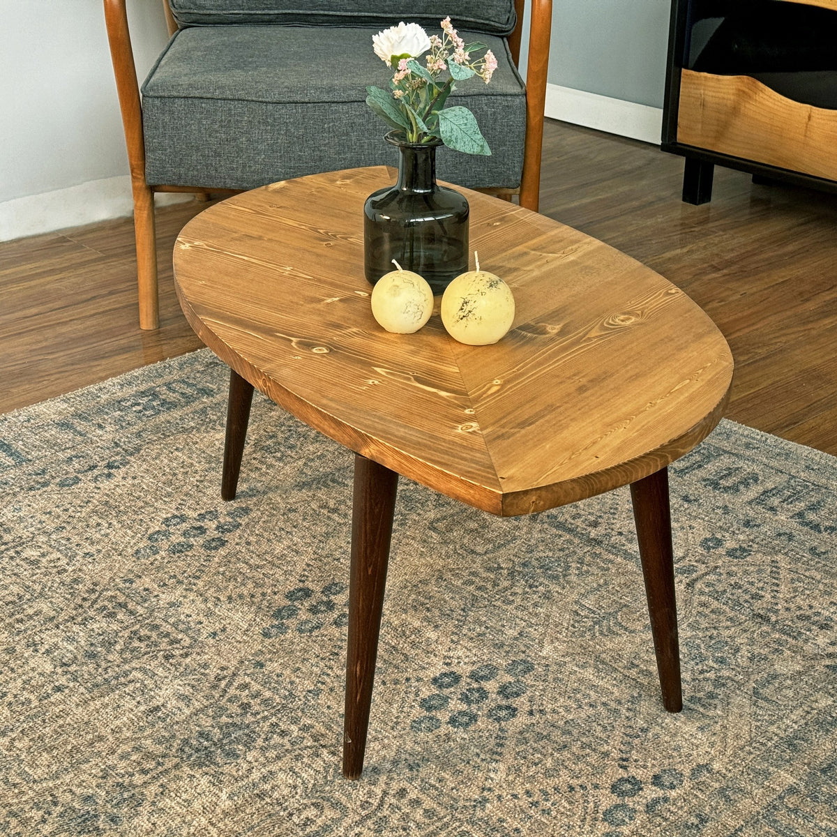 Leaf shaped coffee table with a medium-brown top and dark tapered legs beside a gray armchair