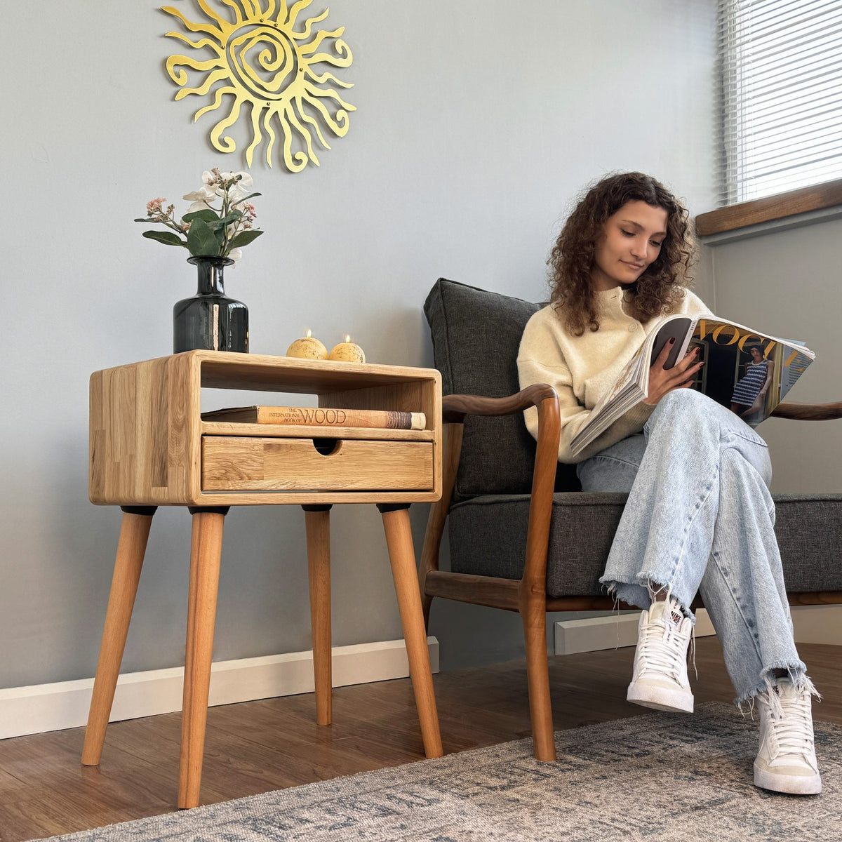 Oak nightstand with drawer beside a gray armchair where a woman reads a magazine, blue wall