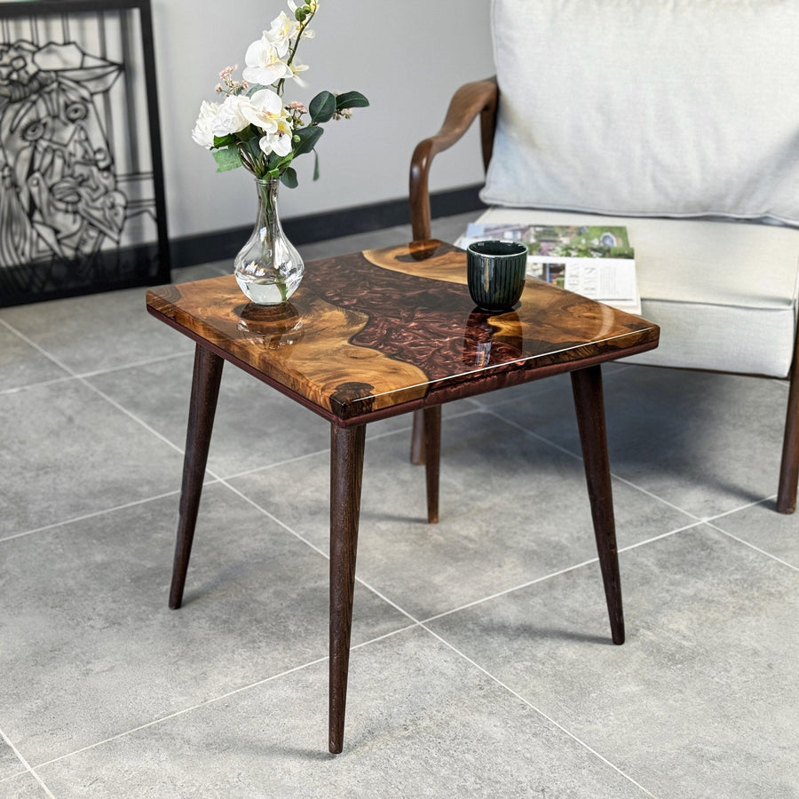 Square walnut side table with a burgundy epoxy river and a vase on top, beside an armchair