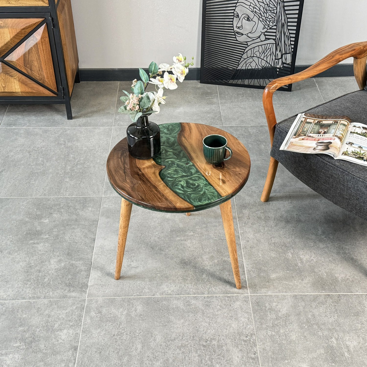 Green resin walnut side table with tapered wooden legs on gray tile, with a sideboard and armchair behind