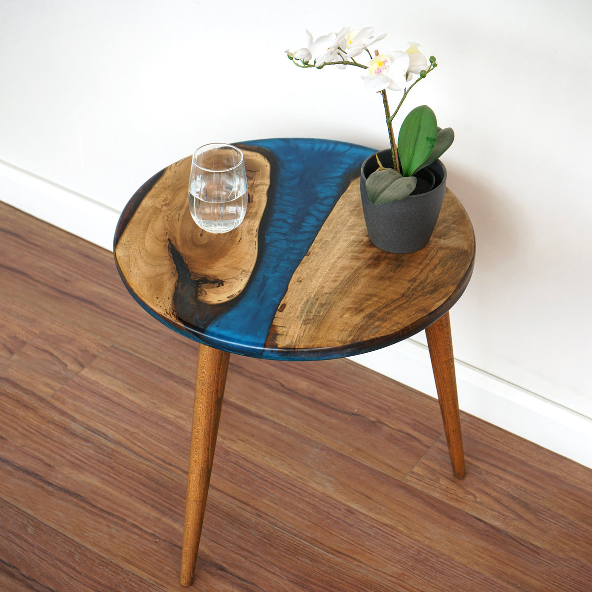 Round live edge walnut side table with a blue epoxy river on tapered legs, on a wood floor