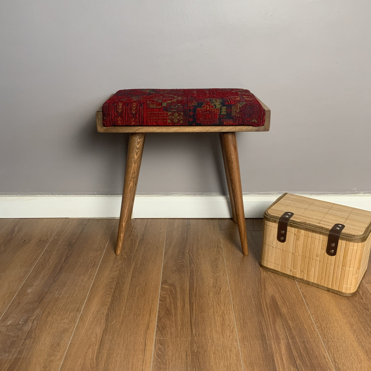 Front view of the wooden piano bench with a red patterned cushion and a wicker box beside it