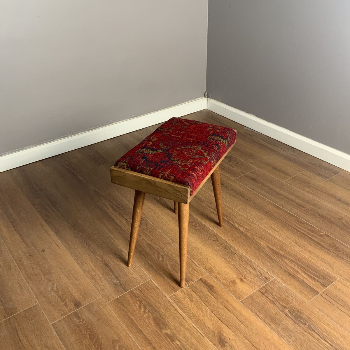 Wooden piano bench with a red patterned cushion seen from above in the corner of a gray room