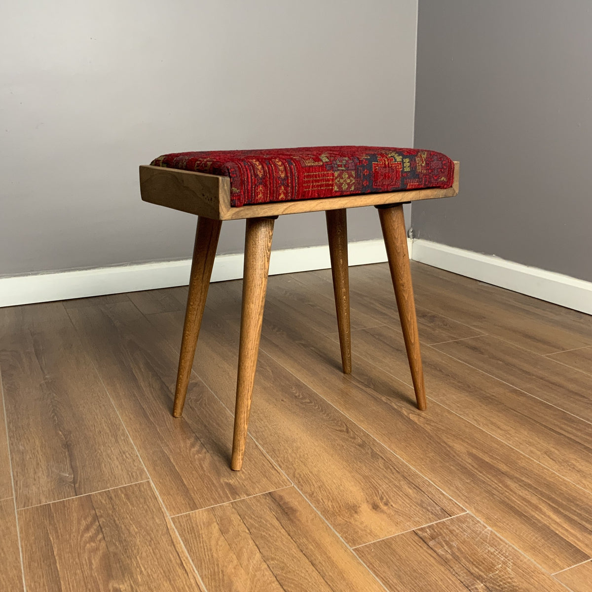 Wooden piano stool with a red patterned cushion on a wood floor in front of a gray wall