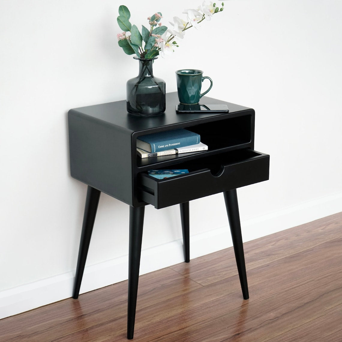 Black mid century nightstand with the drawer pulled half open and books on the shelf, angled view