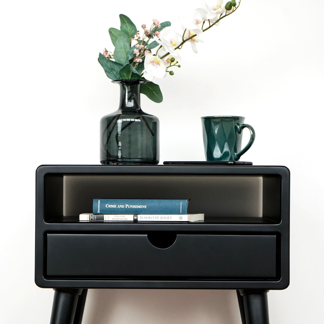 Black bedside table drawer and open shelf framed at the top, with a vase of flowers and a green mug