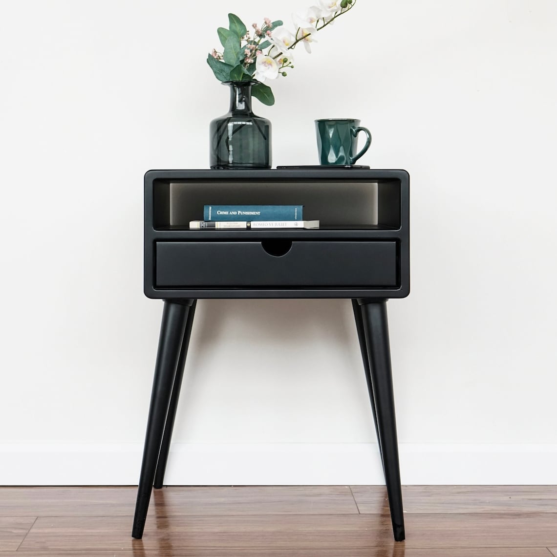 Front view of the black bedside table with an open shelf, books, and a drawer on tapered legs