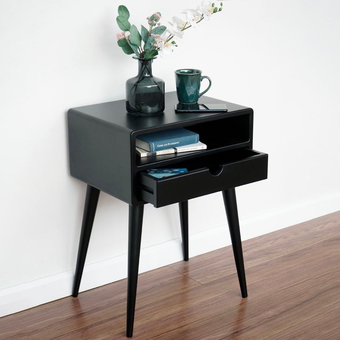 Black mid century nightstand from the front-right with the drawer partly open and books on the shelf
