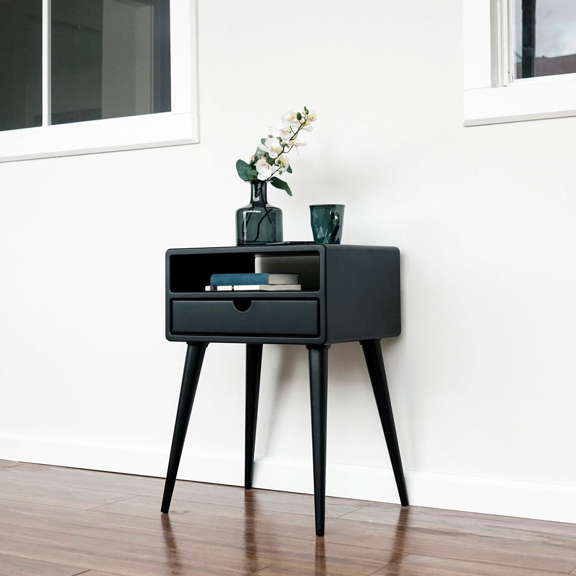 Wide low view of the black nightstand on a wood floor, with a white wall and window behind