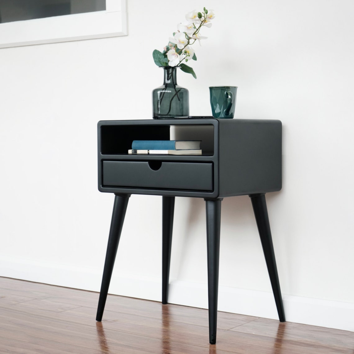 Black nightstand with drawer and shelf, angled view showing the tapered legs and side panel