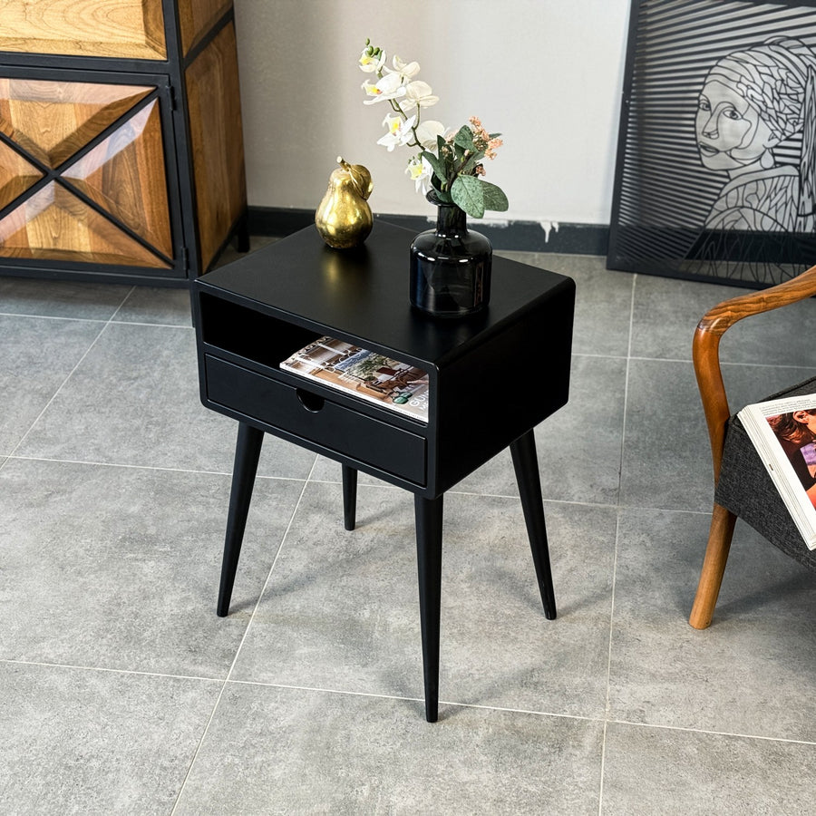 Black mid century side table with an open shelf and drawer on tapered legs, beside an armchair on a gray tile floor