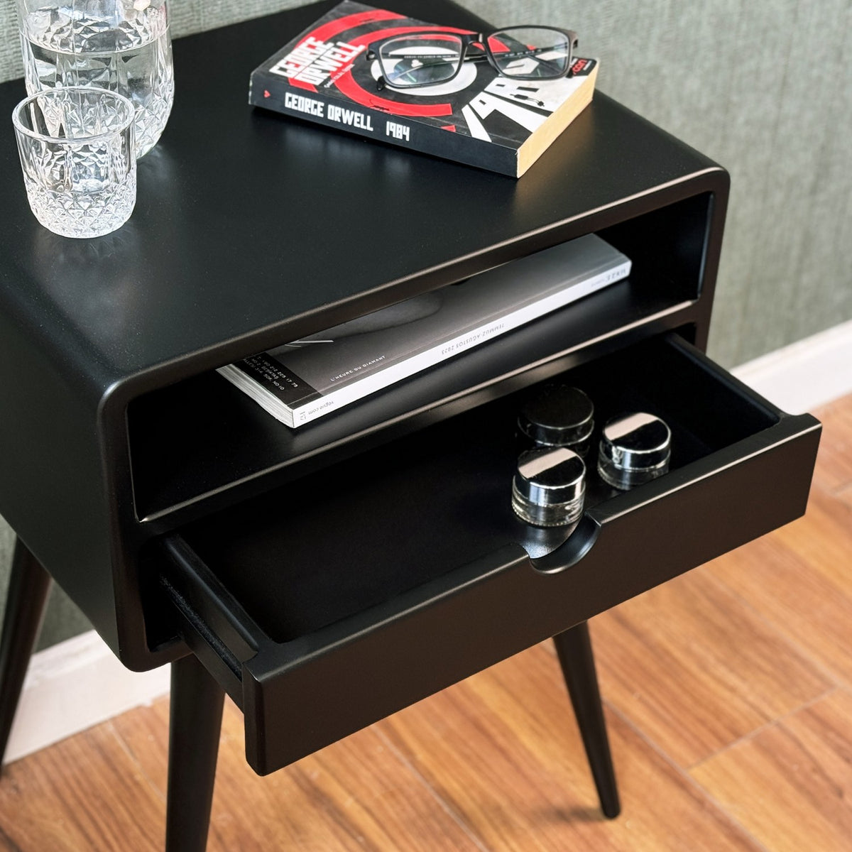 Black side table with the drawer pulled open, a book on top and a rounded shelf above