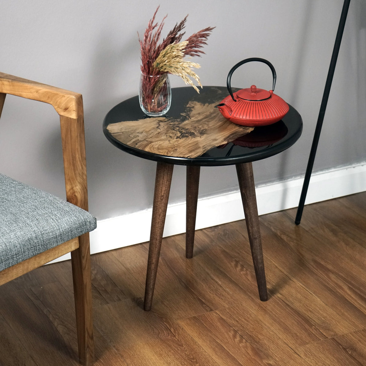 Round black walnut epoxy side table with three tapered legs beside a wooden armchair
