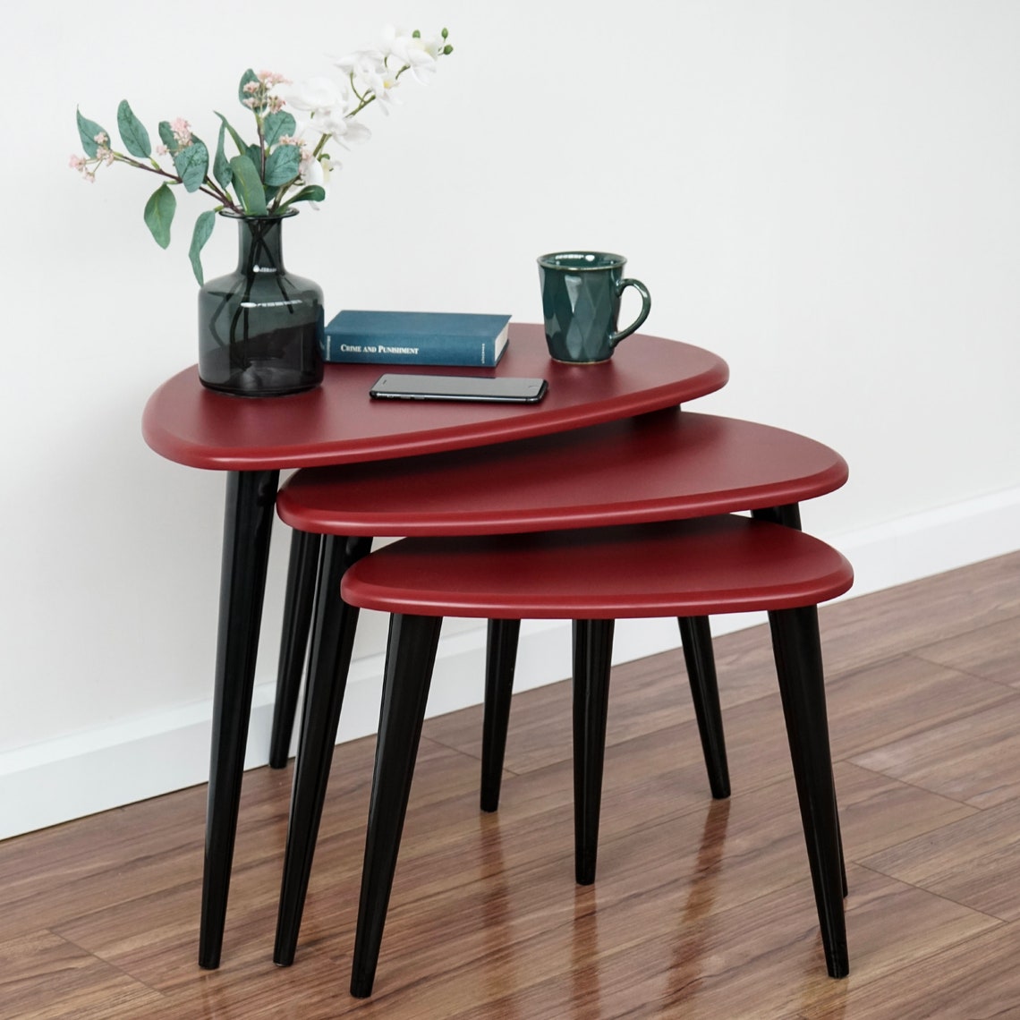 Three burgundy pebble nesting tables with black legs against a white wall on a wood floor