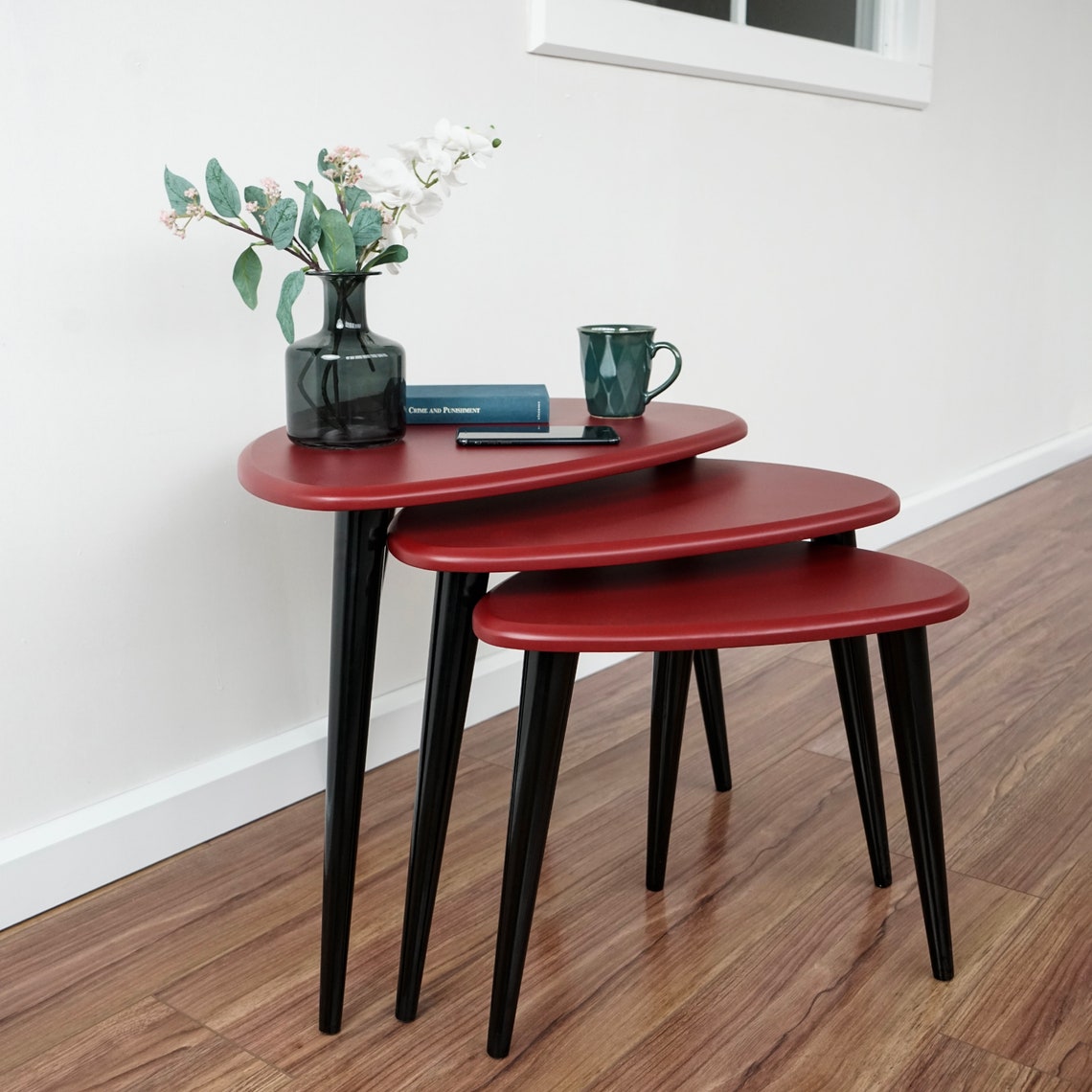 Side view of the burgundy pebble nesting tables set with tapered black legs near a window