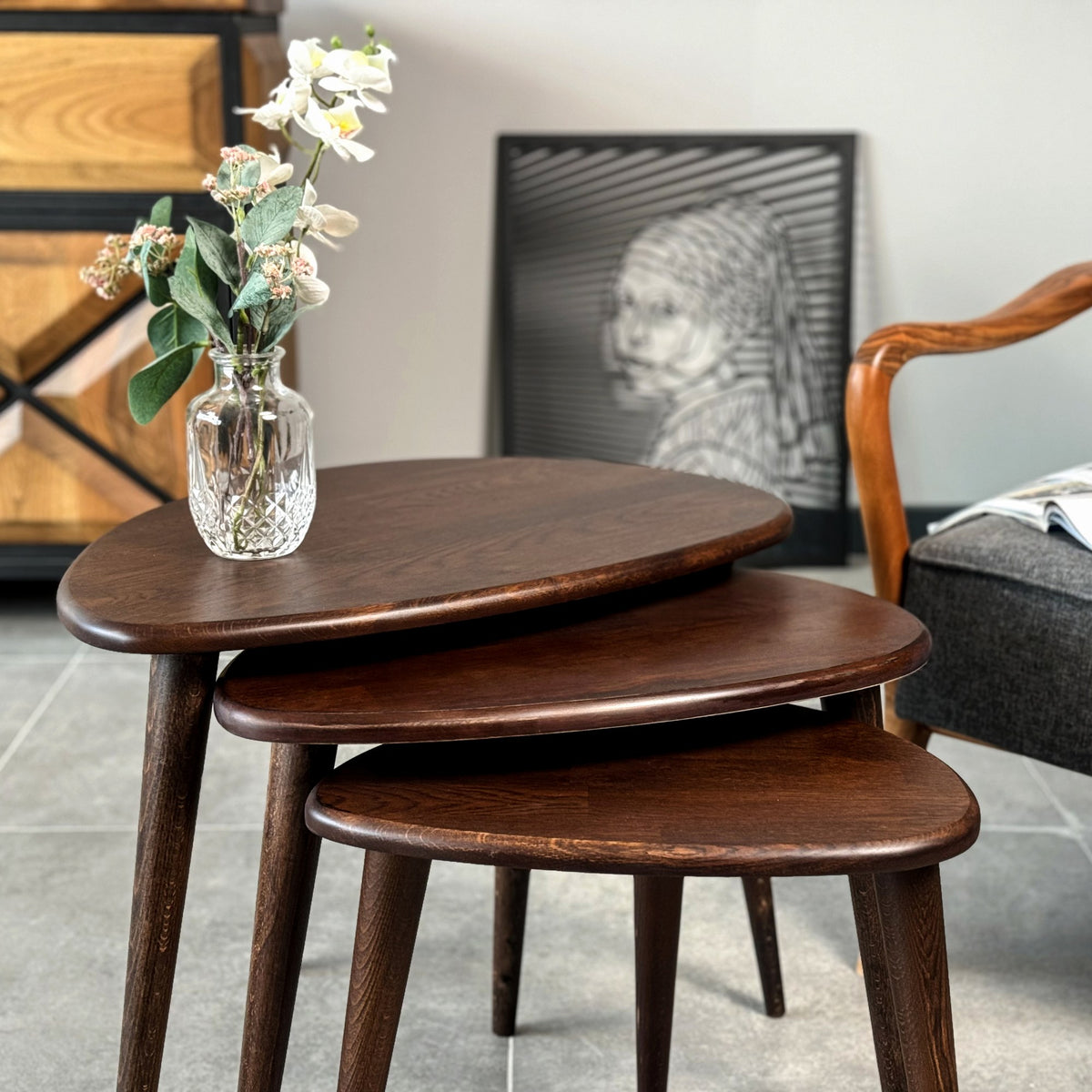 Close view of the chocolate oak nest of tables with pebble-shaped tops, tapered legs and a glass vase