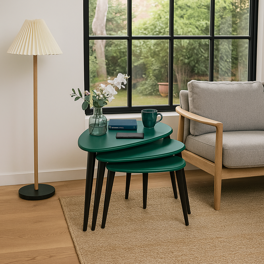 Green Ercol style pebble nest of three tables with black legs beside an armchair and a window