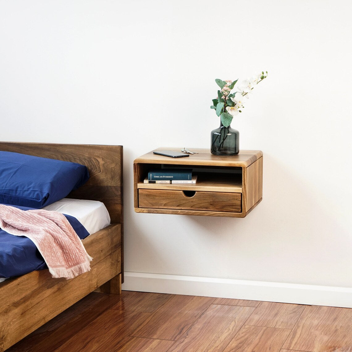 Walnut floating nightstand with shelf and drawer next to a bed with blue bedding, wide crop with floor