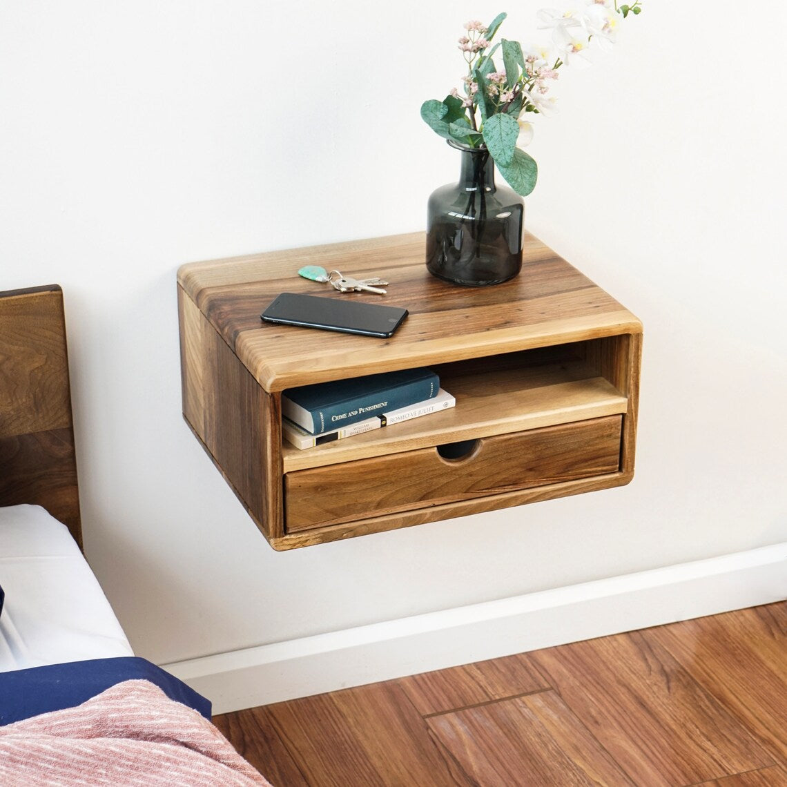 Floating walnut nightstand with open shelf and drawer, top-angle view beside a bed headboard