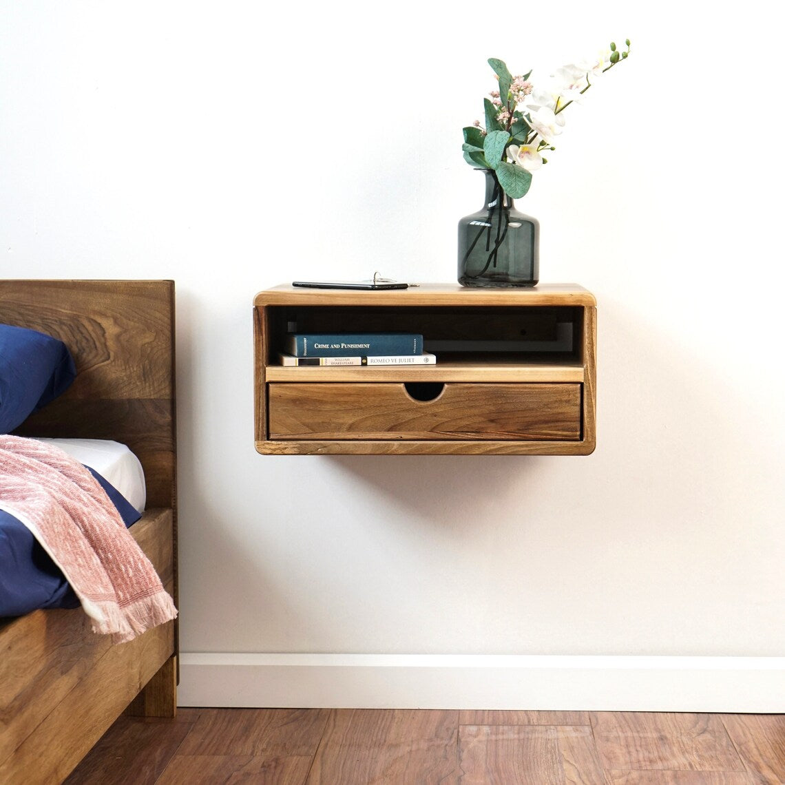 Small floating walnut nightstand with open shelf and drawer on a white wall above a wood floor
