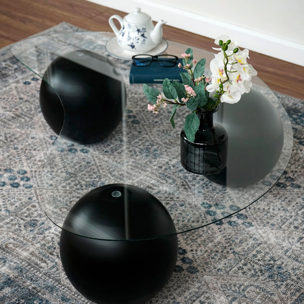 High angle of the glass coffee table with black ball legs on a gray patterned rug, tea set on top