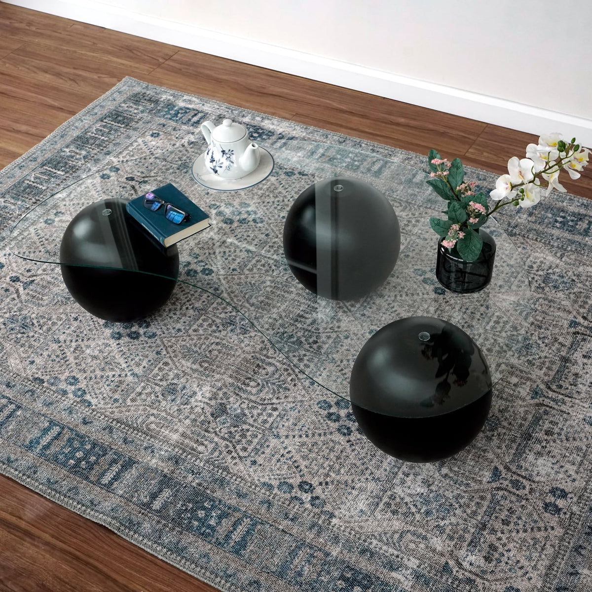Glass coffee table with three black ball legs from a high side angle on a patterned rug